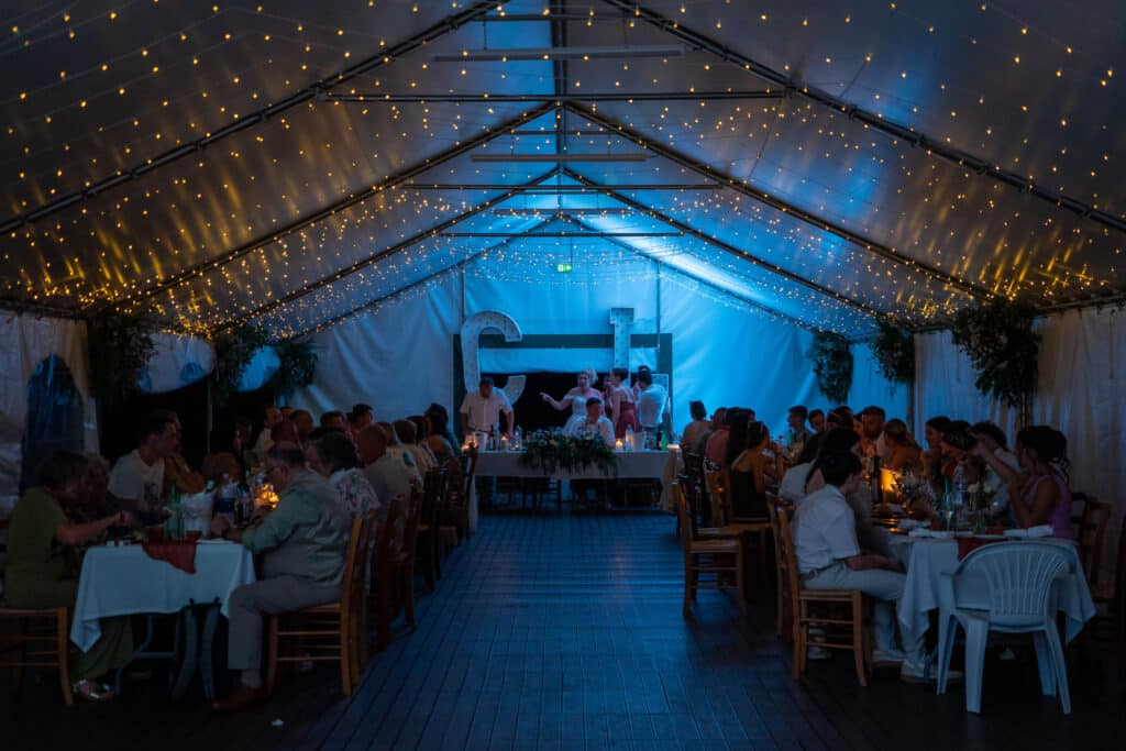 Fête de mariage sous tente, ambiance tamisée avec lumières jaunes et éclairage bleu, invités attablés.