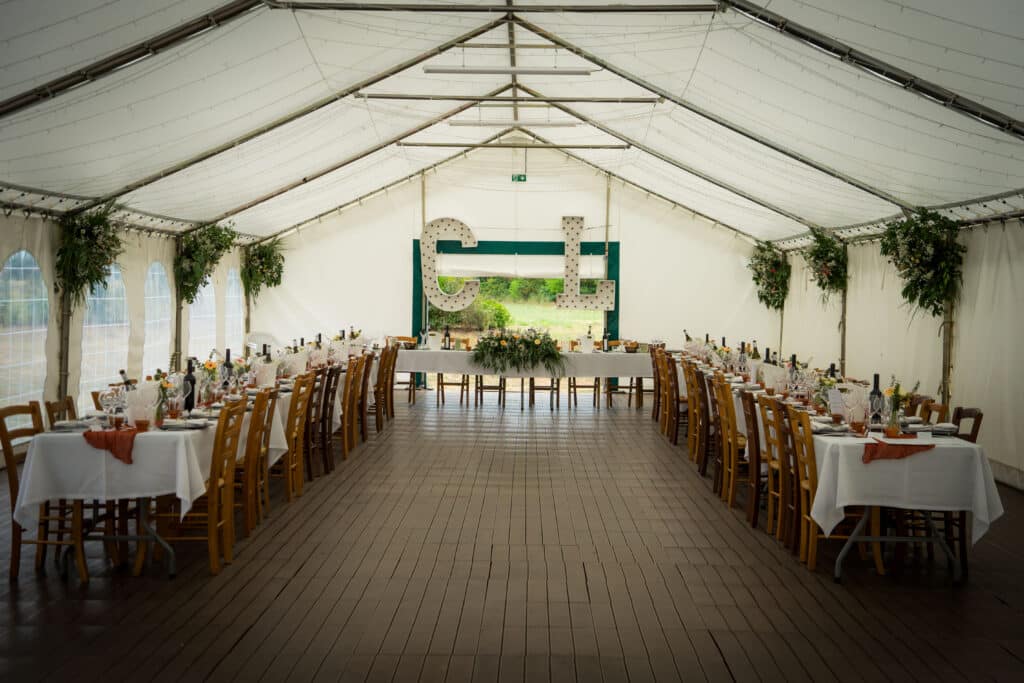 Tente de réception décorée pour un mariage, tables dressées avec vaisselle et vin, grandes lettres 'C' et 'L' lumineuses à l'arrière.